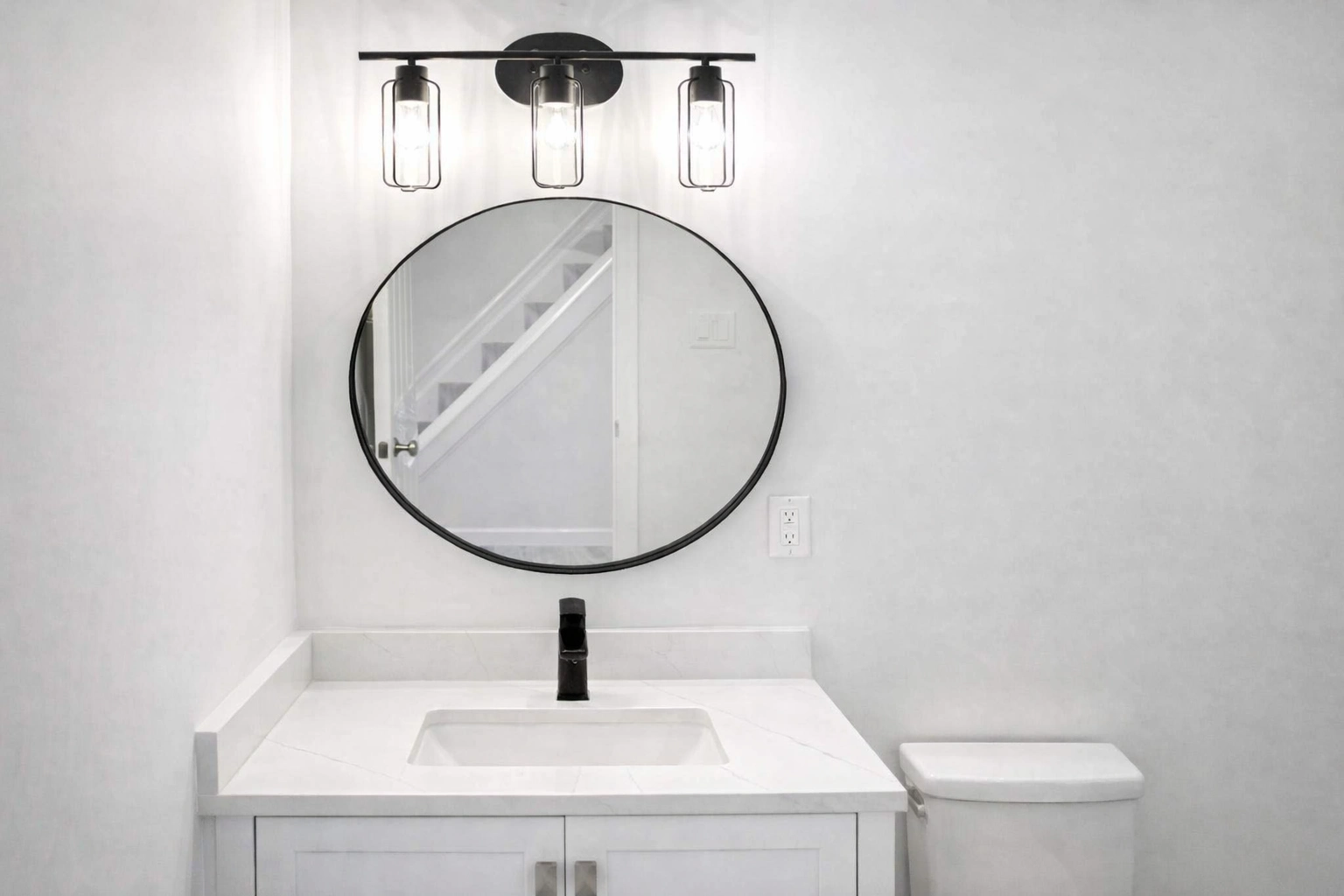 Modern bathroom remodeler in Neptune City, NJ showcasing a white shaker vanity, integrated sink, black faucet, and round wall mirror with industrial-style lighting.