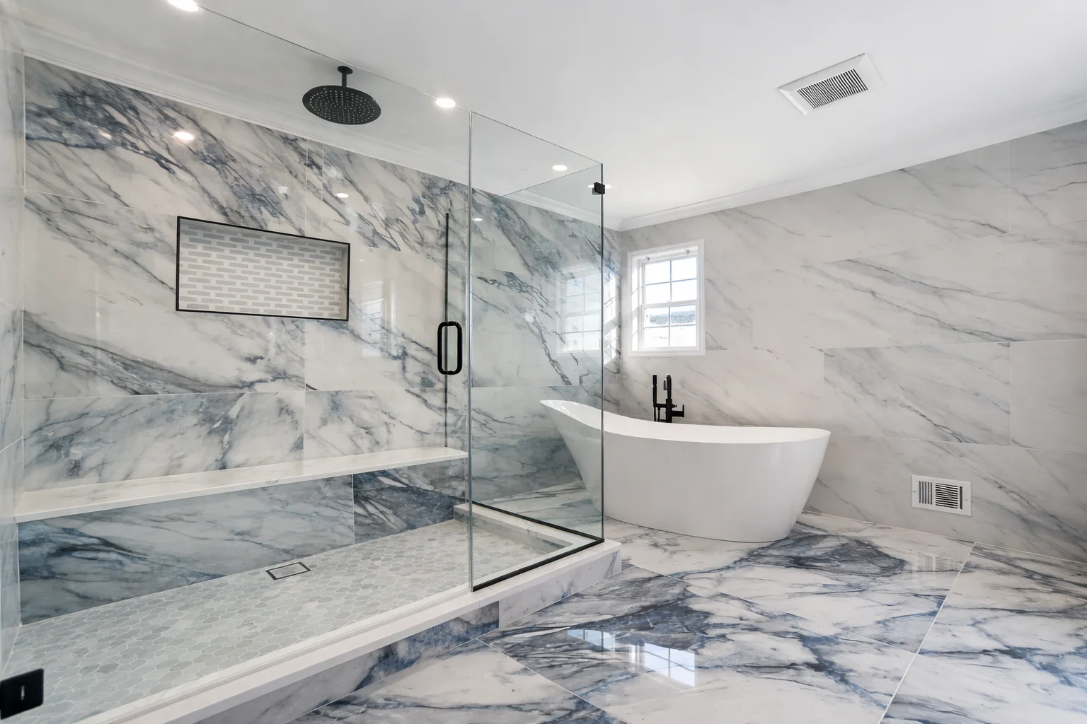 Wide view of a luxury bathroom remodeling project in Marlboro, NJ featuring a glass-enclosed walk-in shower, freestanding soaking tub, and marble tile floors and walls with modern black fixtures.