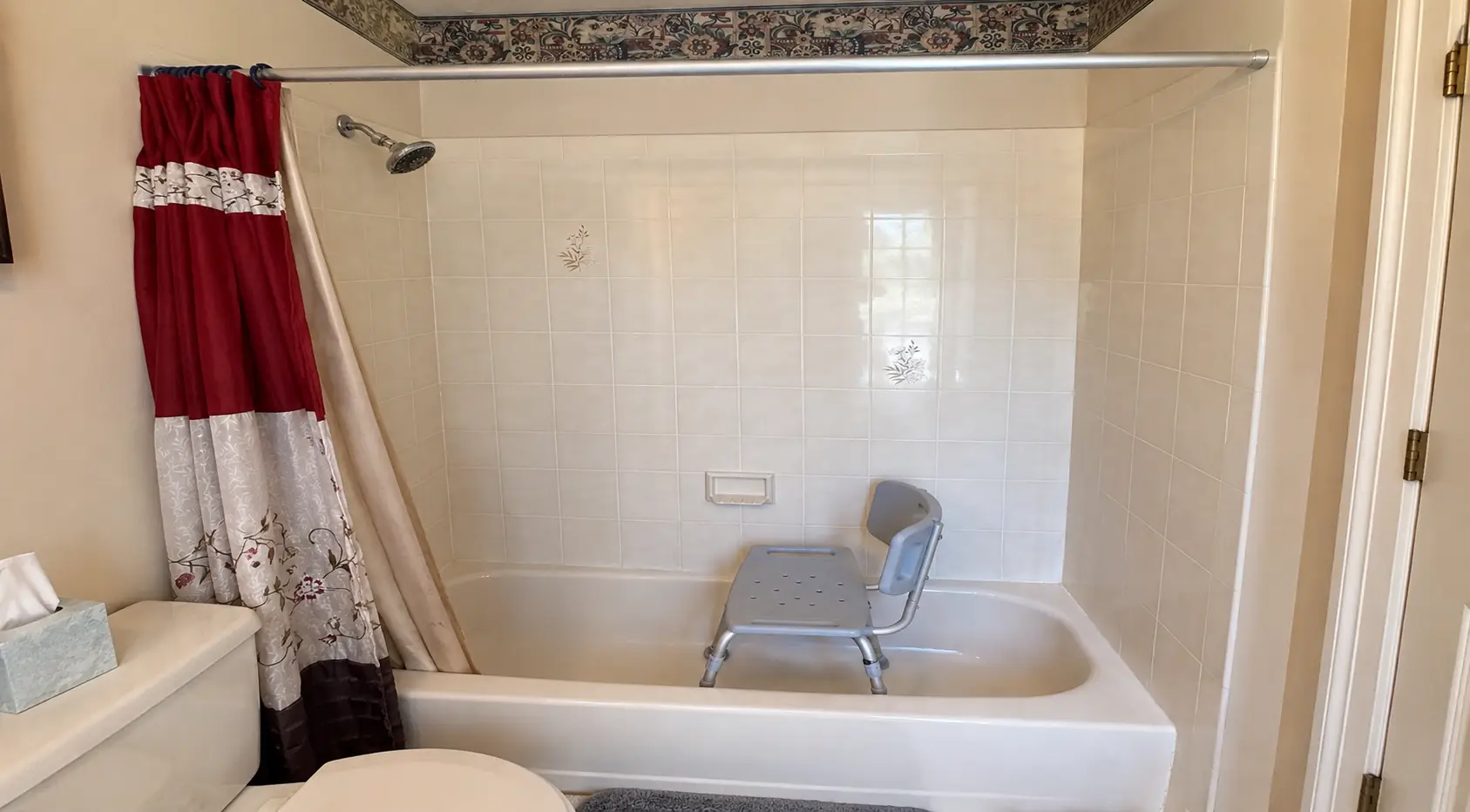 Before photo of an outdated bathtub with tiled shower walls, shower curtain, and bath chair prior to a tub-to-shower conversion.