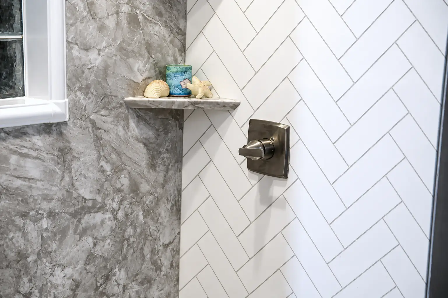 Remodeled shower with white herringbone tile, gray marble-look wall panels, brushed nickel shower trim, and corner shelf.